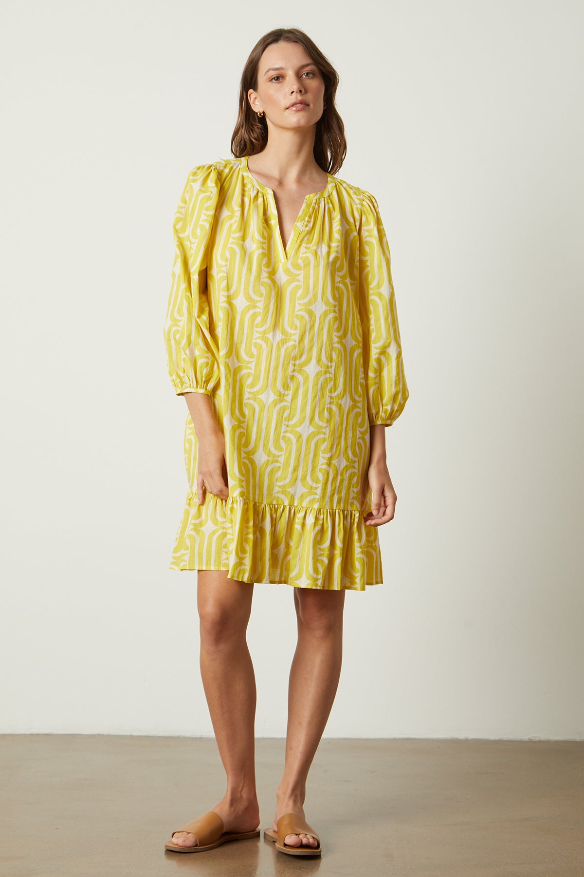 A woman wearing a yellow FELICITY PRINTED BOHO DRESS by Velvet by Graham & Spencer with a ruffled sleeve and a mod-inspired print.-26235561017537