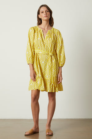A woman wearing a yellow FELICITY PRINTED BOHO DRESS by Velvet by Graham & Spencer with mod-inspired print and sandals.