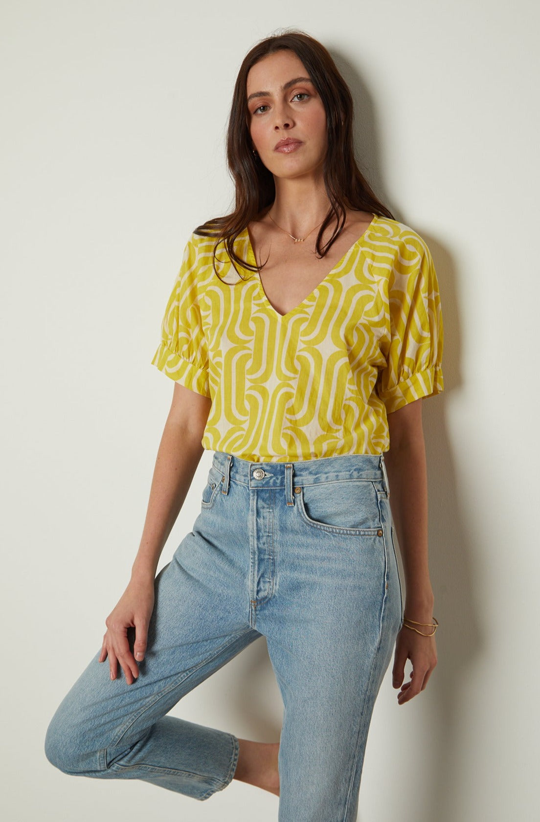 a woman in a yellow Velvet by Graham & Spencer JODY PRINTED TOP and jeans leaning against a wall.-26142636409025
