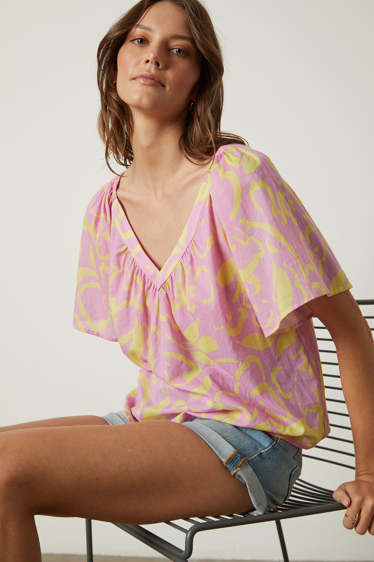 A woman is sitting on a chair wearing a Velvet by Graham & Spencer Liliana Printed V-Neck Top in pink and yellow.-25954331328705