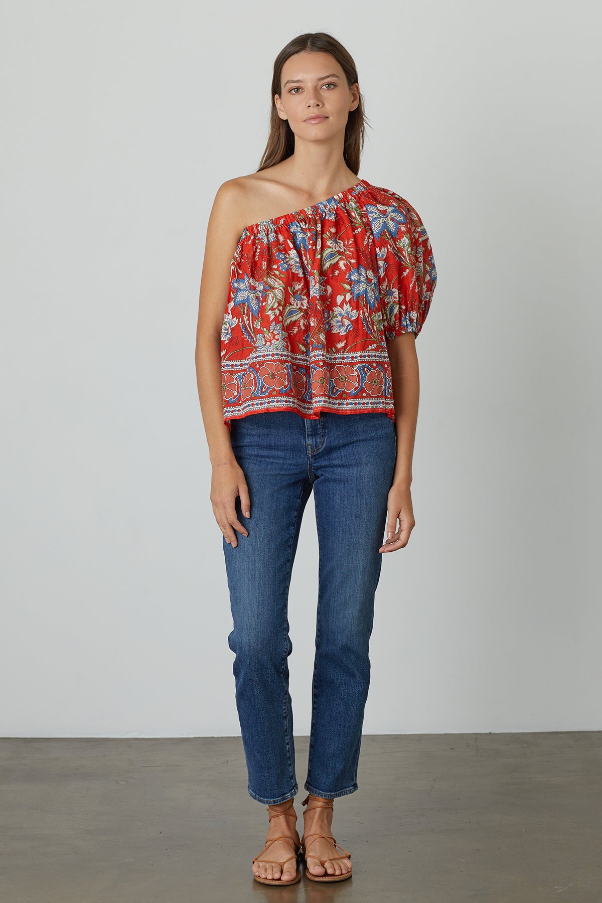   A woman stands against a plain background, wearing the DIANA ONE SHOULDER PRINTED TOP by Velvet by Graham & Spencer, paired with blue jeans and sandals. 