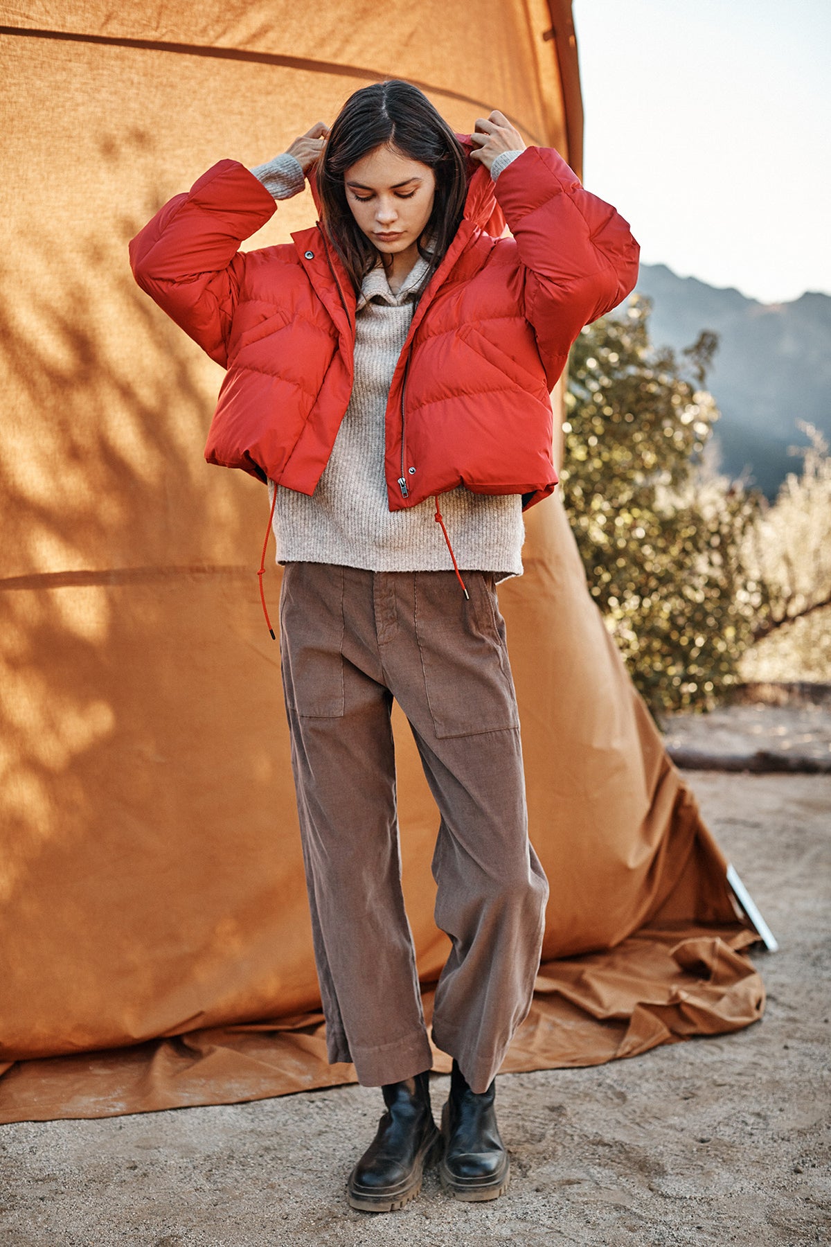 A person stands outdoors beside a tent, wearing a bright red RAYLIN CROPPED PUFFER JACKET by Velvet by Graham & Spencer, a cozy beige sweater, brown pants, and black boots. The background features a natural landscape with trees and mountains.-23195050410177