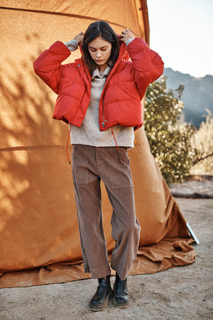 A person stands outdoors beside a tent, wearing a bright red RAYLIN CROPPED PUFFER JACKET by Velvet by Graham & Spencer, a cozy beige sweater, brown pants, and black boots. The background features a natural landscape with trees and mountains.