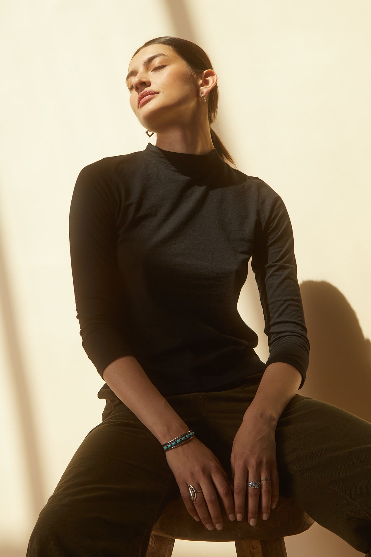 A woman sitting on a stool in a QUINNY 3/4 SLEEVE MOCK NECK TEE by Velvet by Graham & Spencer, a perfect layering piece made of soft cotton knit.-25175811686593