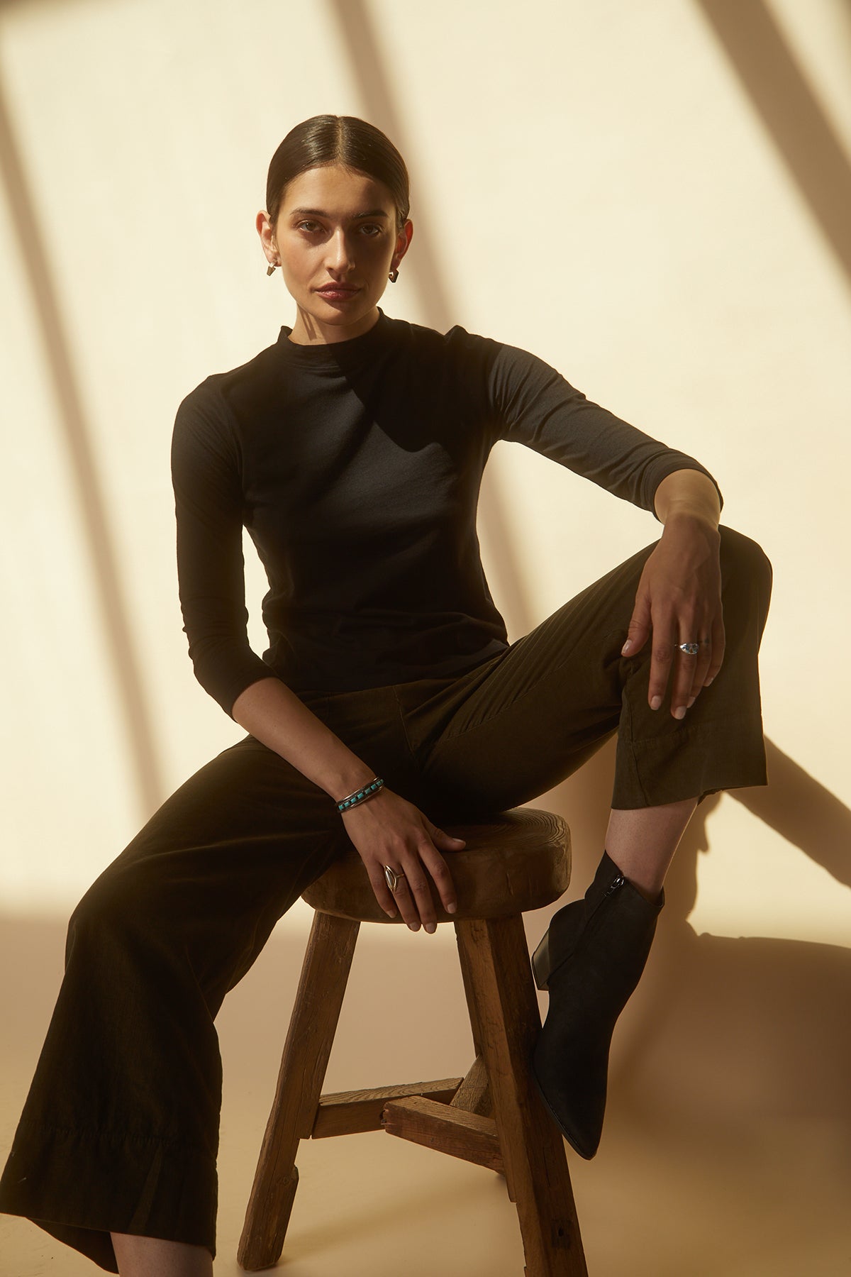 A woman sitting on a stool in a QUINNY 3/4 SLEEVE MOCK NECK TEE by Velvet by Graham & Spencer, wearing wide leg pants.-25175811719361