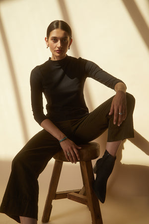 A woman sitting on a stool in a QUINNY 3/4 SLEEVE MOCK NECK TEE by Velvet by Graham & Spencer, wearing wide leg pants.