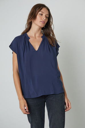 The model is wearing a SARENA CAP SLEEVE BLOUSE by Velvet by Graham & Spencer, a blue top with a v-neck and cap sleeves.