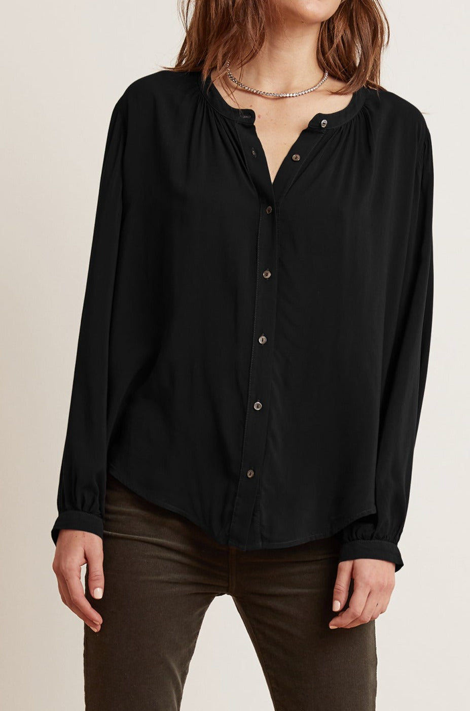   Person wearing a traditional SERAFINA BUTTON-UP BLOUSE by Velvet by Graham & Spencer in black with long sleeves and olive green pants, standing with hands relaxed by their sides—perfectly capturing that work-to-evening transition. 