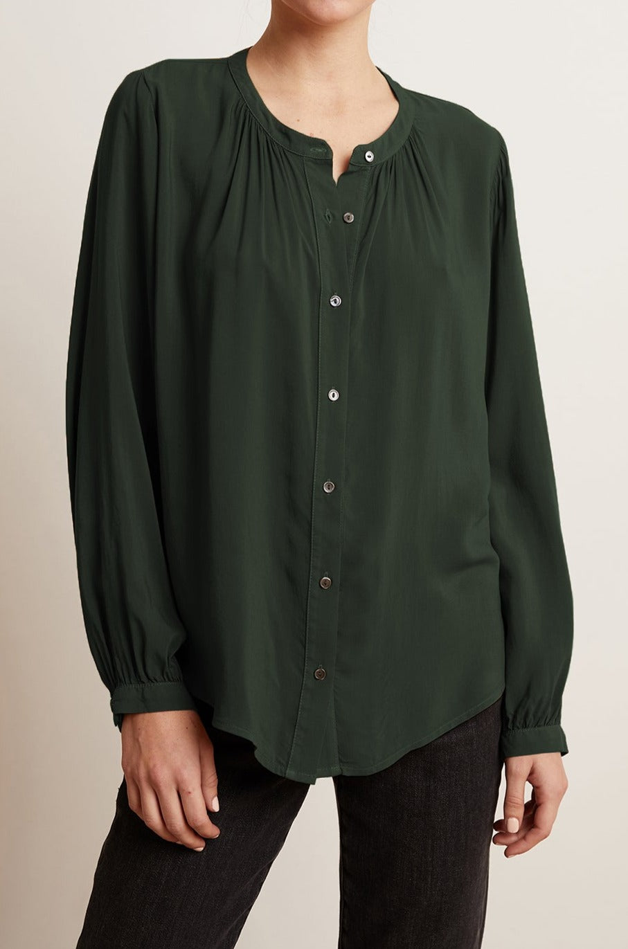   A person wearing the SERAFINA BUTTON-UP BLOUSE in dark green from Velvet by Graham & Spencer, along with black pants, stands in front of a plain light background. The blouse features long sleeves and a round neck, making it perfect for work-to-evening transitions. 