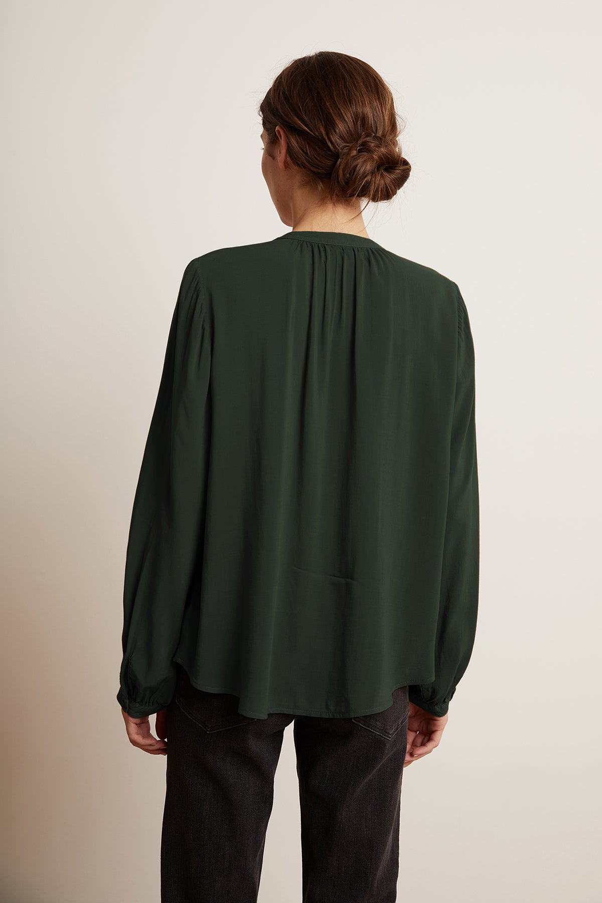 A person with brown hair tied up in a bun is facing away from the camera, wearing the Velvet by Graham & Spencer SERAFINA BUTTON-UP BLOUSE, a long-sleeved dark green top that effortlessly transitions from work to evening, paired with dark pants.-8165405458513