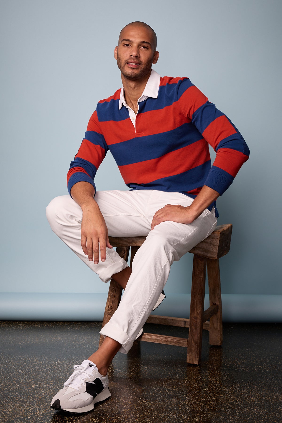   Model sitting on wooden stool wearing Pierre Rugby Stripe long sleeve polol with broad blue and red stripes and white collar with white denim and New Balance sneakers full length front 