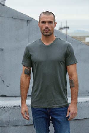 A man with a shaved head stands outdoors, wearing a green SAMSEN WHISPER CLASSIC V-NECK TEE by Velvet by Graham & Spencer and jeans. He has tattoos on his arms and is facing the camera against an urban rooftop background.