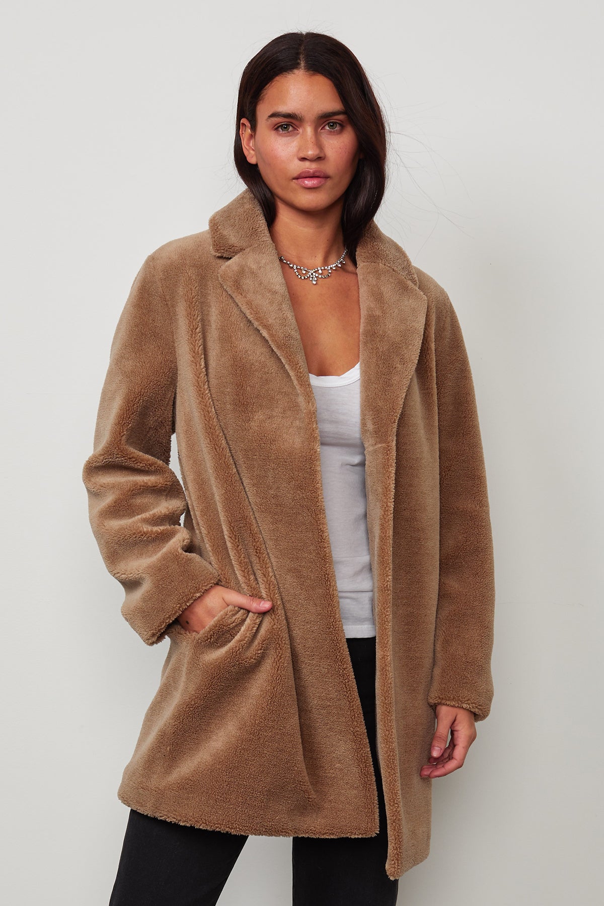   A person stands against a neutral background wearing the TRISHELLE LUX SHERPA OVERSIZED COAT by Velvet by Graham & Spencer, with giant popper buttons over a white top and black pants. 