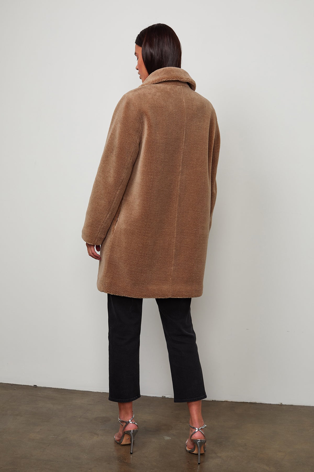 A person stands facing away, wearing a brown, oversized **TRISHELLE LUX SHERPA OVERSIZED COAT** by **Velvet by Graham & Spencer** with giant popper buttons and a high collar, black pants, and heeled sandals on a plain background.-8031344328785