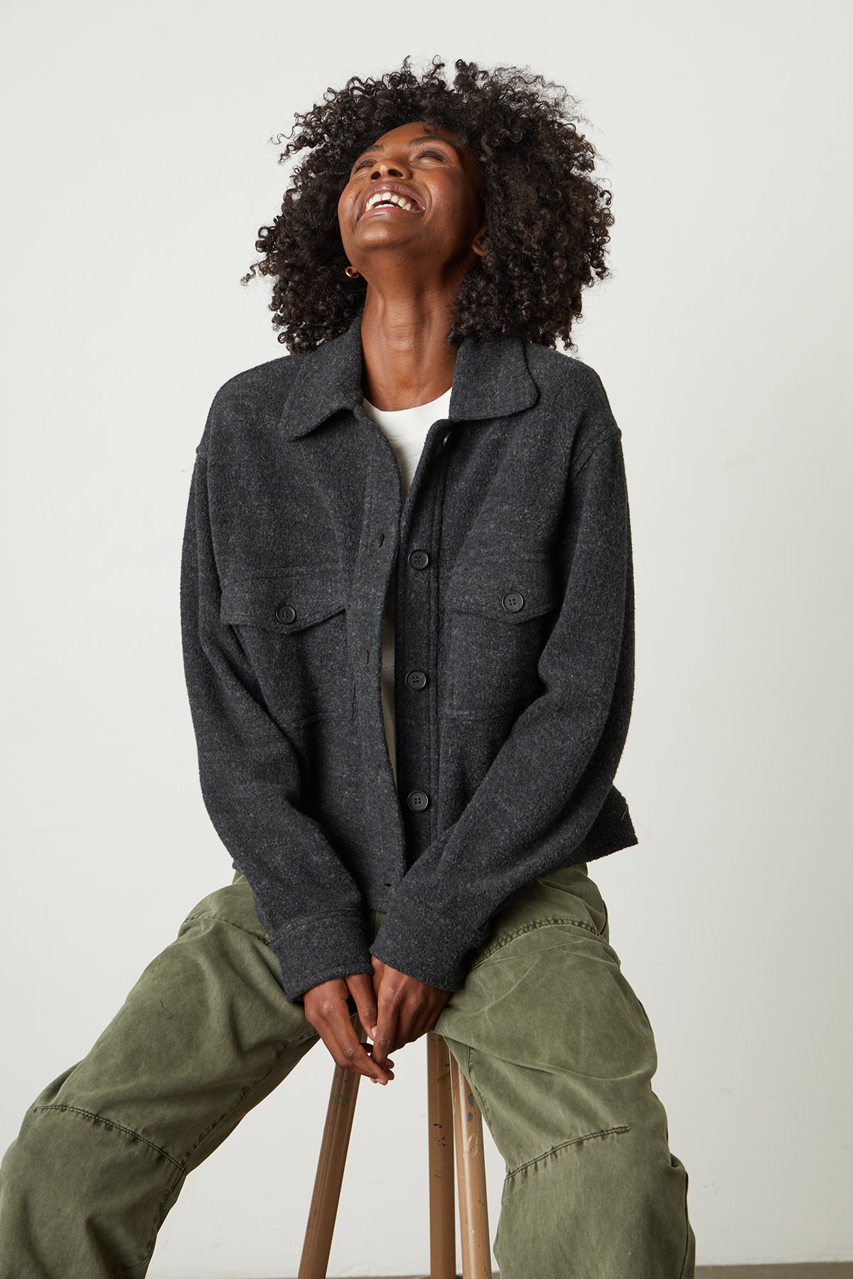 A woman is sitting on a stool wearing the Velvet by Graham & Spencer KAMERON FLEECE SHACKET and green pants.-26068045332673