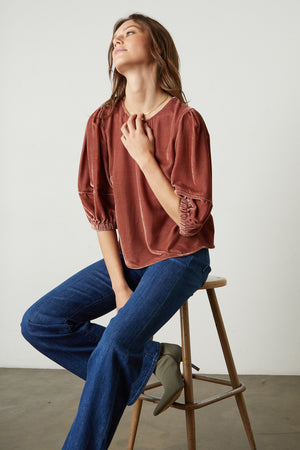 A woman is sitting on a stool wearing jeans and a Nancy Silk Velvet Puff Sleeve Top by Velvet by Graham & Spencer.