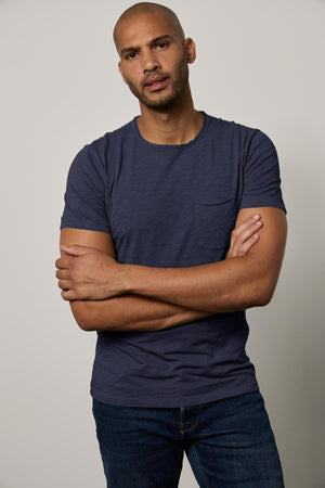 A bald man wearing a blue Velvet by Graham & Spencer CHAD TEE with raw-edge details, standing with his arms crossed, looking directly at the camera against a light gray background.