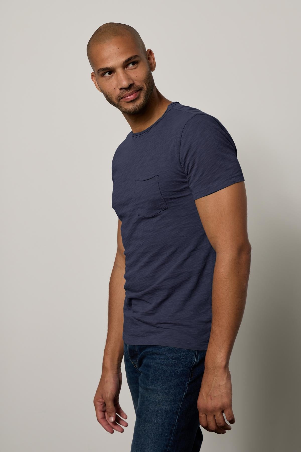 A man in a blue CHAD TEE from Velvet by Graham & Spencer with raw-edge details and jeans standing with his body turned slightly, looking at the camera with a slight smile.-26146501263553
