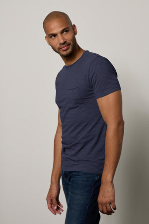 A man in a blue CHAD TEE from Velvet by Graham & Spencer with raw-edge details and jeans standing with his body turned slightly, looking at the camera with a slight smile.