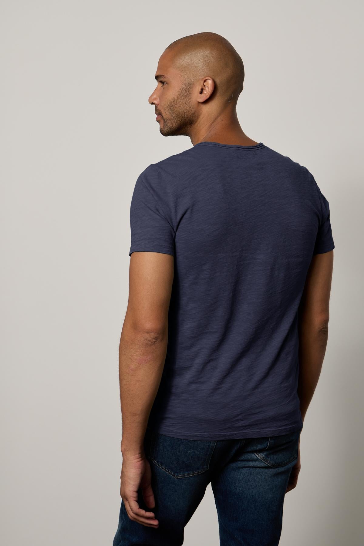   A bald man in a CHAD TEE from Velvet by Graham & Spencer with raw-edge details and jeans stands turned to the side against a neutral background. 