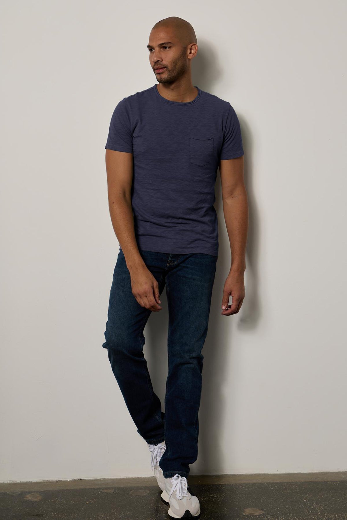 A man stands leaning against a wall, wearing a dark blue textured cotton slub CHAD TEE by Velvet by Graham & Spencer, jeans, and white sneakers. He is bald and gazing to his left.-26146501230785