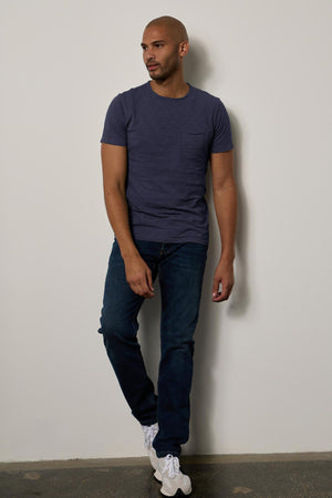 A man stands leaning against a wall, wearing a dark blue textured cotton slub CHAD TEE by Velvet by Graham & Spencer, jeans, and white sneakers. He is bald and gazing to his left.