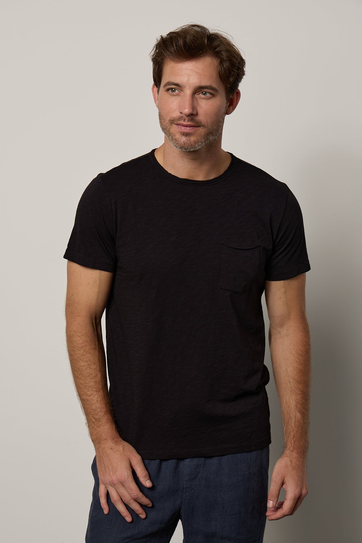A man in a Velvet by Graham & Spencer CHAD TEE with raw-edge details and blue jeans stands against a neutral background, looking slightly to his left.-26098082382017