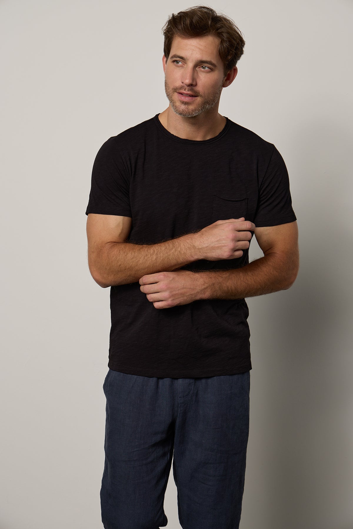 Man in a Velvet by Graham & Spencer California classic dark CHAD TEE and blue pants standing with arms crossed, looking to the side against a neutral background.-26098082513089