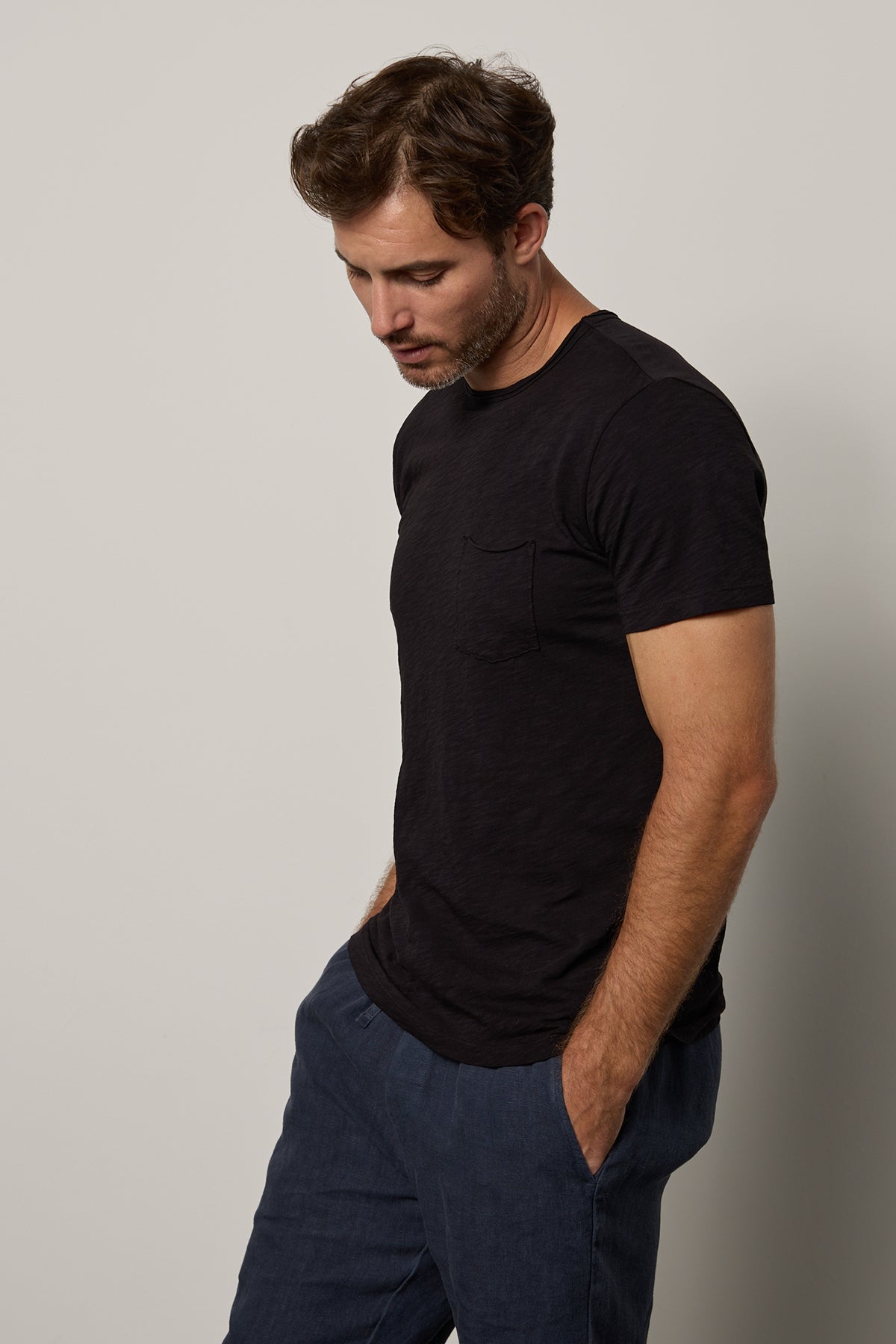 A man in a Velvet by Graham & Spencer CHAD TEE with raw-edge details and jeans looking downward while standing against a neutral background.-26098082447553