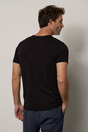 A man standing with his back turned, wearing a black Velvet by Graham & Spencer CHAD TEE with raw-edge details and blue jeans, looking to his right side against a neutral background.