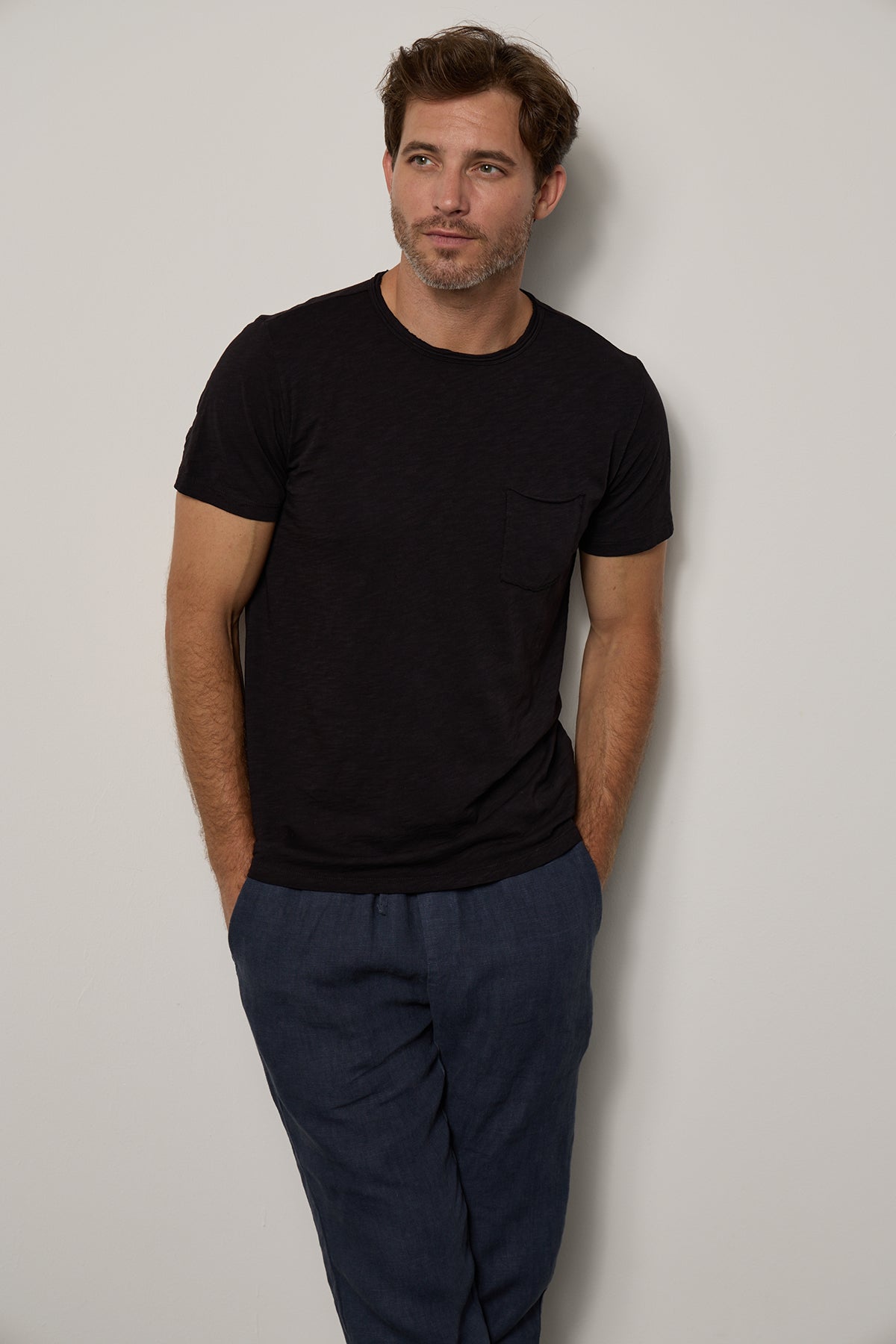   Man in a black textured cotton slub CHAD TEE by Velvet by Graham & Spencer and navy pants standing against a neutral background, looking to his left with a subtle smile. 