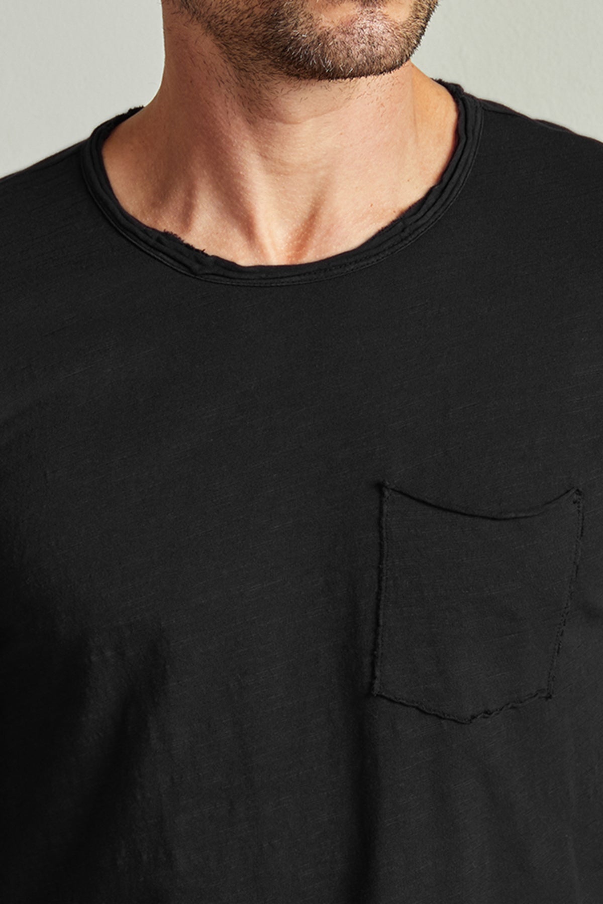  Close-up of a man wearing a black Velvet by Graham & Spencer CHAD TEE with raw-edge details and a pocket, focusing on the shirt and neck area. 