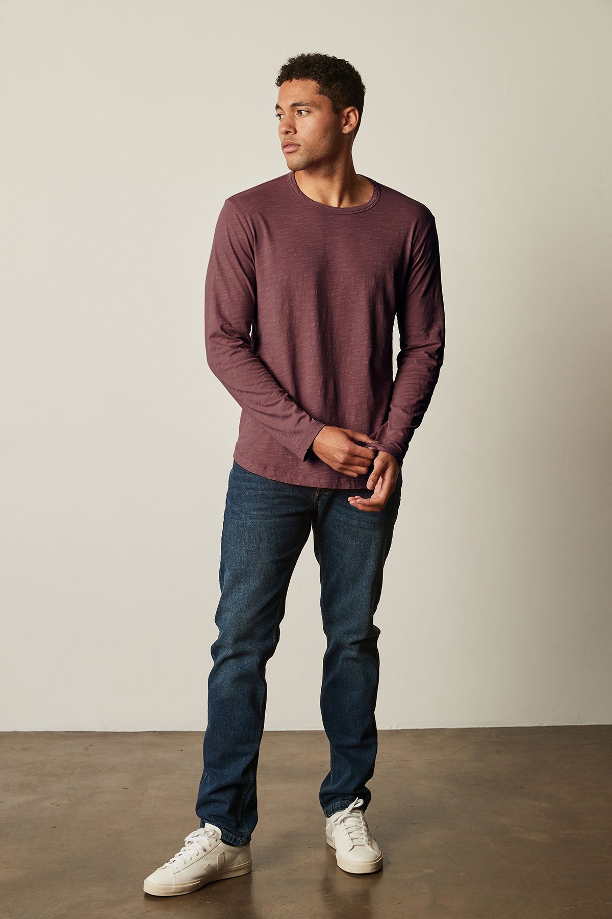 A man wearing a maroon KAI TEE with a crew neckline and long sleeves from Velvet by Graham & Spencer, blue jeans, and white sneakers stands against a plain background. He is looking to his right with his hands clasped in front of him.-25793842577601