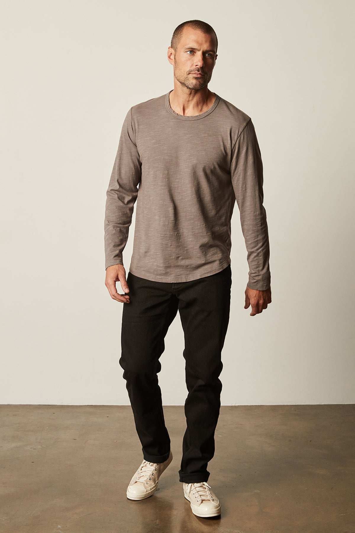   A man stands against a plain background, wearing a long-sleeved brown KAI TEE by Velvet by Graham & Spencer with a crew neckline, black pants, and white sneakers. 