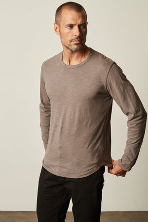 A man with short hair wearing a long-sleeved, crew neckline gray KAI TEE from Velvet by Graham & Spencer and black pants stands against a plain background, looking to the side with one hand in his pocket.