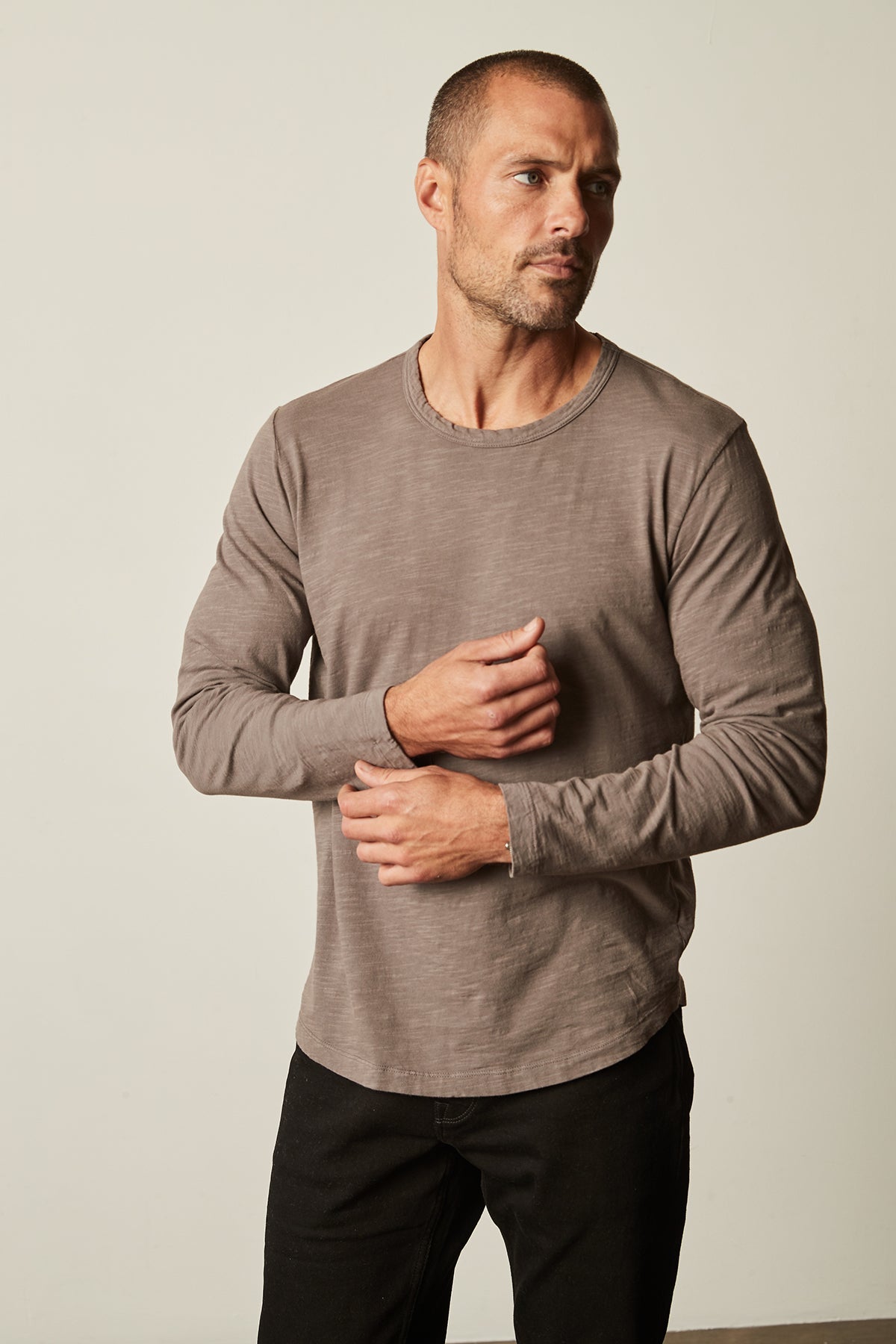   A man with short hair wearing a long-sleeve grey KAI TEE by Velvet by Graham & Spencer and black pants stands against a plain background, looking to his right and adjusting his sleeve. 