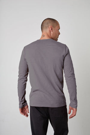 A person with a shaved head, wearing a long-sleeved gray SIMEON RAW EDGE COTTON SLUB TEE by Velvet by Graham & Spencer and dark pants, is shown from the back. There is a small tattoo on the back of their neck.
