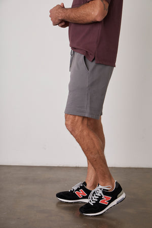 A man wearing Velvet by Graham & Spencer JAXSON DRAWSTRING SHORT and sneakers standing in front of a white wall.