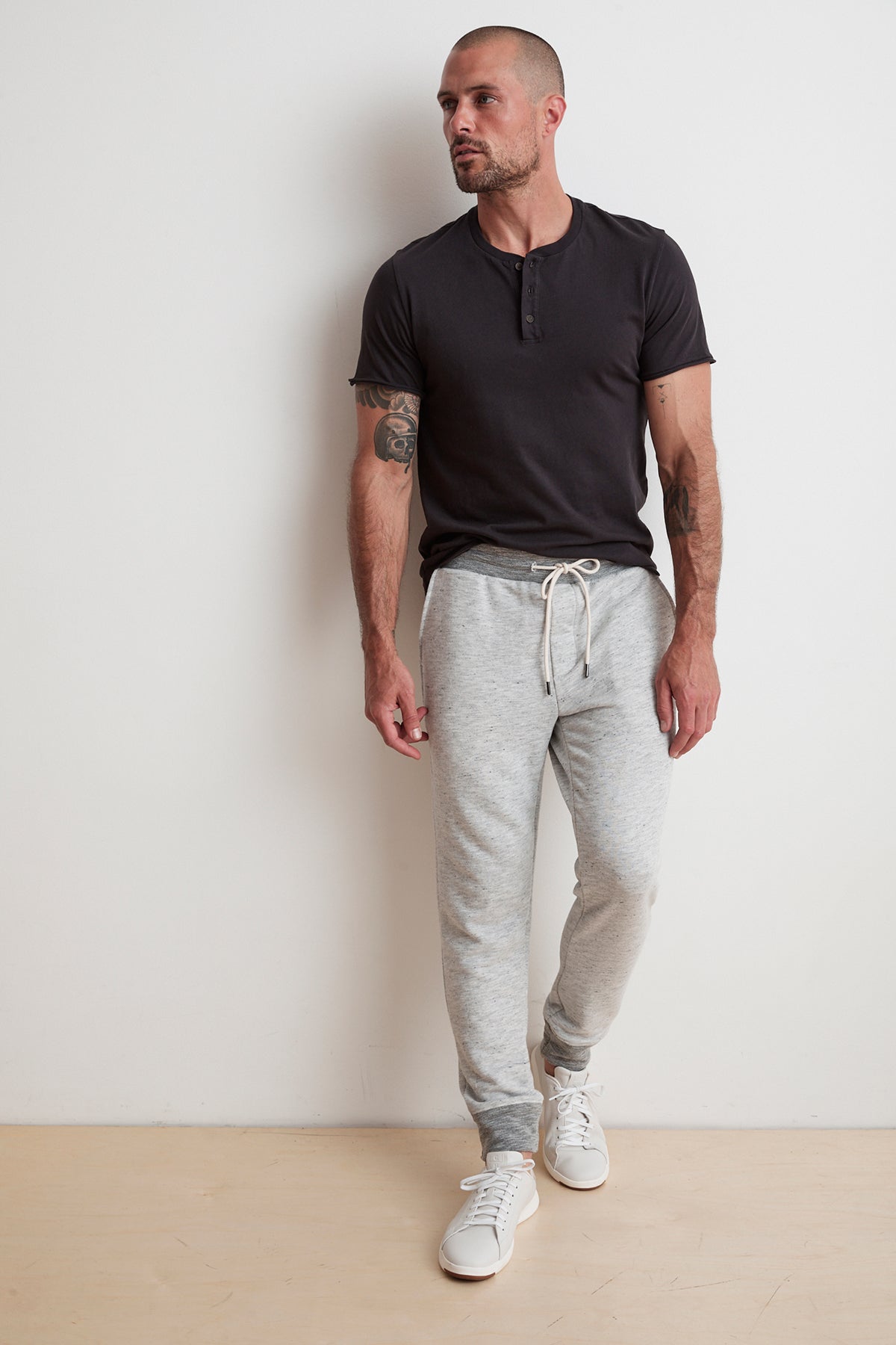 A man wearing a black henley shirt and light gray BOBBY FRENCH TERRY JOGGER pants with an elastic waistband from Velvet by Graham & Spencer stands against a plain white wall. He has tattoos on his arms and is looking to his left.-6344103657553