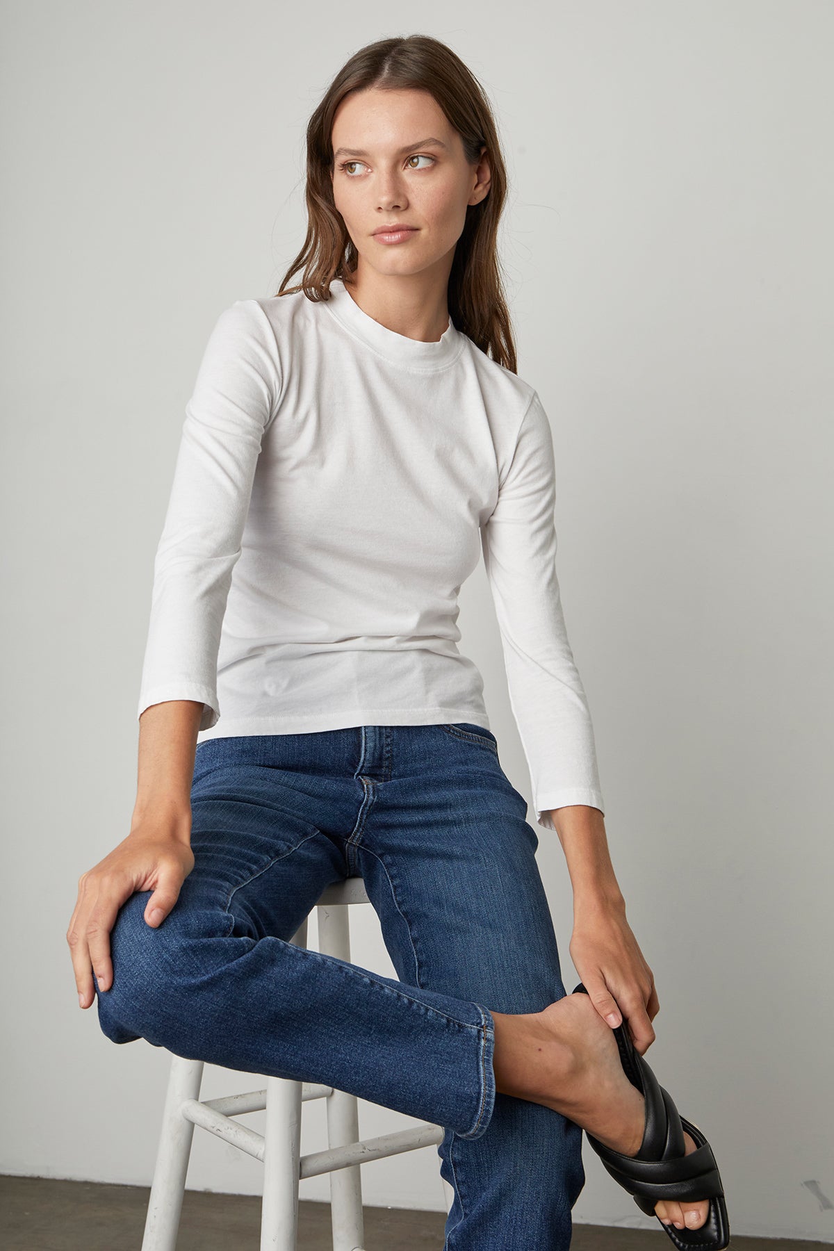   Model sitting on stool wearing Quinny 3/4 Sleeve Mock Neck Tee in white with blue denim and black sandals 