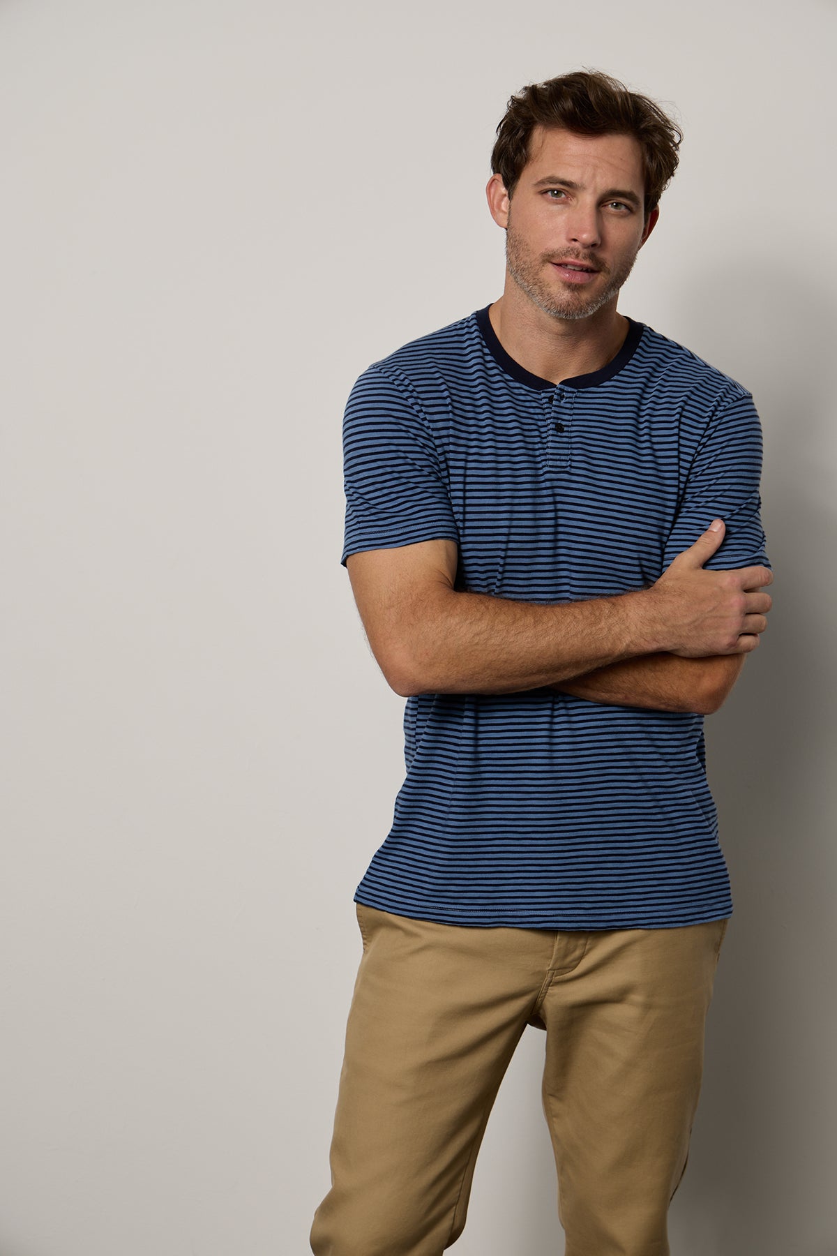 Male model standing with arms folded wearing Albert Henley with medium and dark blue stripes and Aiden pant in khaki-26249474605249