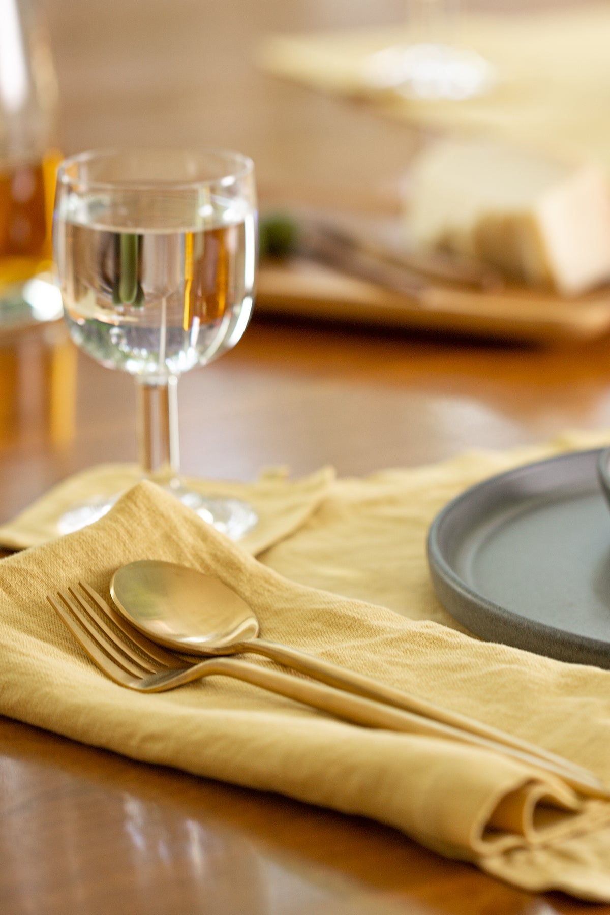  A neatly set dining table with an elegant yellow Jenny Graham Home linen napkin, a spoon, and a glass of water, with blurry plates and a meal in the background. 