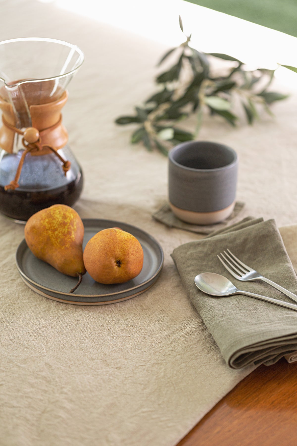 An elegant table setting featuring two pears on a plate, a coffee carafe, a cup, and neatly arranged silverware on Jenny Graham Home linen napkins.-15227668693185