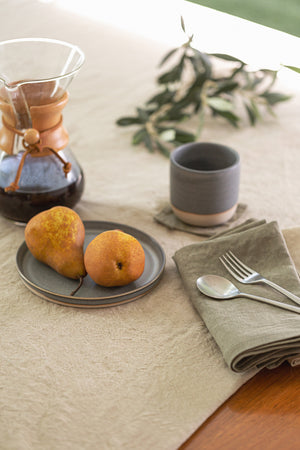 An elegant table setting featuring two pears on a plate, a coffee carafe, a cup, and neatly arranged silverware on Jenny Graham Home linen napkins.