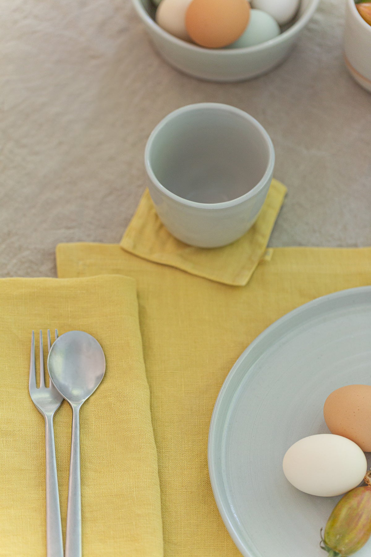 A breakfast table setting with a pale blue plate, silver fork and spoon on an elegant yellow Jenny Graham Home linen napkin, and a cup surrounded by eggs in bowls.-14899237224641