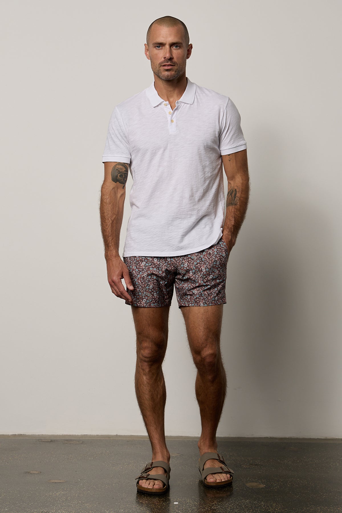   A man in a white shirt and RICARDO PRINTED SWIM SHORTS by Velvet by Graham & Spencer is standing in front of a white wall. 