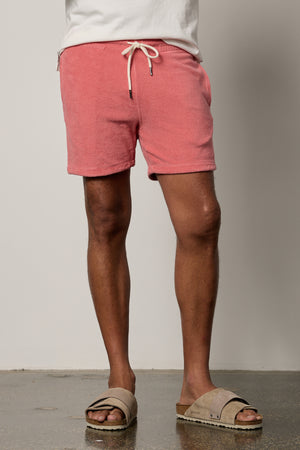 A man wearing OZZIE TERRY SHORT by Velvet by Graham & Spencer shorts and sandals is standing in front of a white wall.
