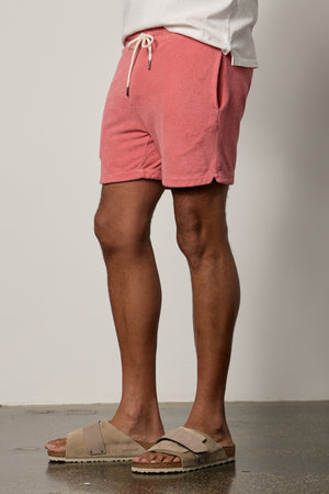 A man in OZZIE TERRY SHORT by Velvet by Graham & Spencer and sandals standing in front of a wall.