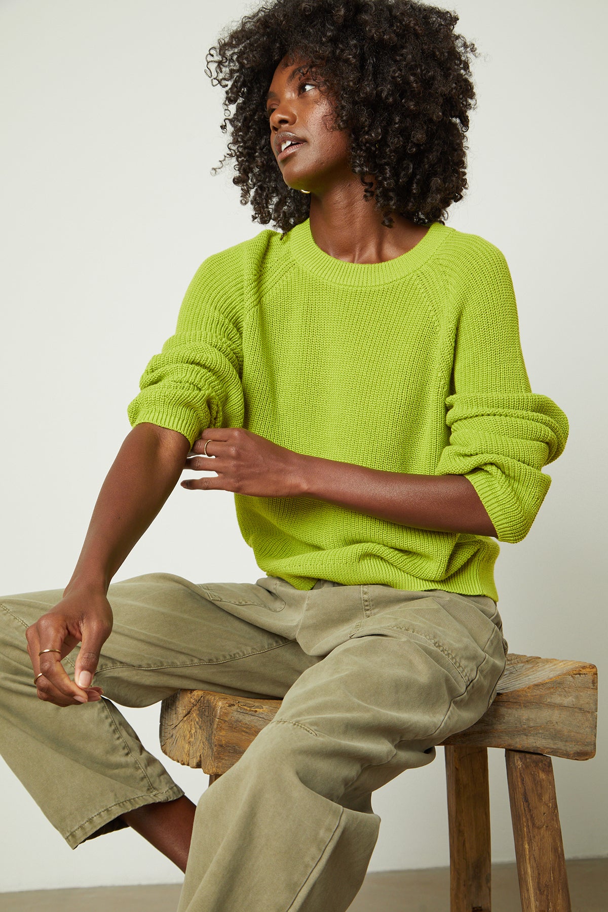   A woman is sitting on a stool in a Velvet by Graham & Spencer LINAH CREW NECK SWEATER in lime green. 