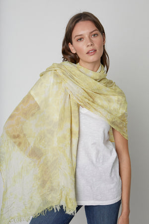 A woman wearing a white t-shirt and blue jeans drapes the super soft knit, light yellow CHLOE TIE DYE SCARF by Velvet by Graham & Spencer over her shoulders. She has long brown hair and is standing against a plain, light gray background.